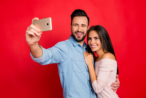 Portrait of young, caucasian, attractive, lovely, cute, smiling, positive, couple, man making selfie with his wonderful woman on smart phone over red background, 14 february