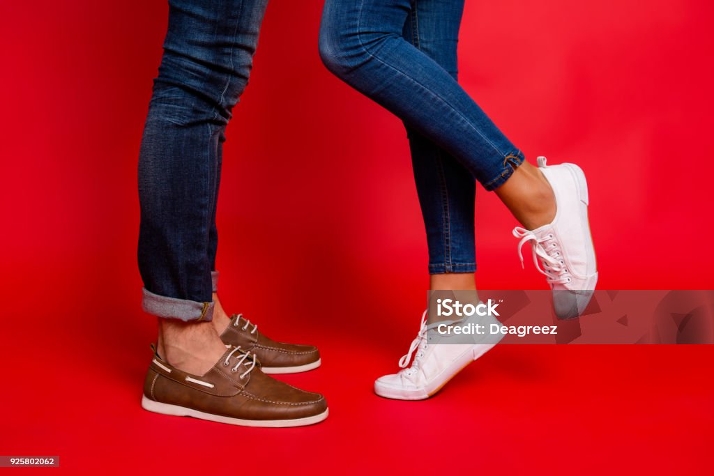 Closeup photo of woman and man legs in jeans, pants and shoes, girl with raised leg, stylish couple kissing during date, isolated over red background, he vs she Shoe Stock Photo