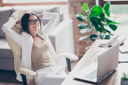Nice, independent, cute woman in white suit, formal wear, glasses sitting on chair at her desk in office, holding arms behind the head with close eyes, thinking about holidays, vacation
