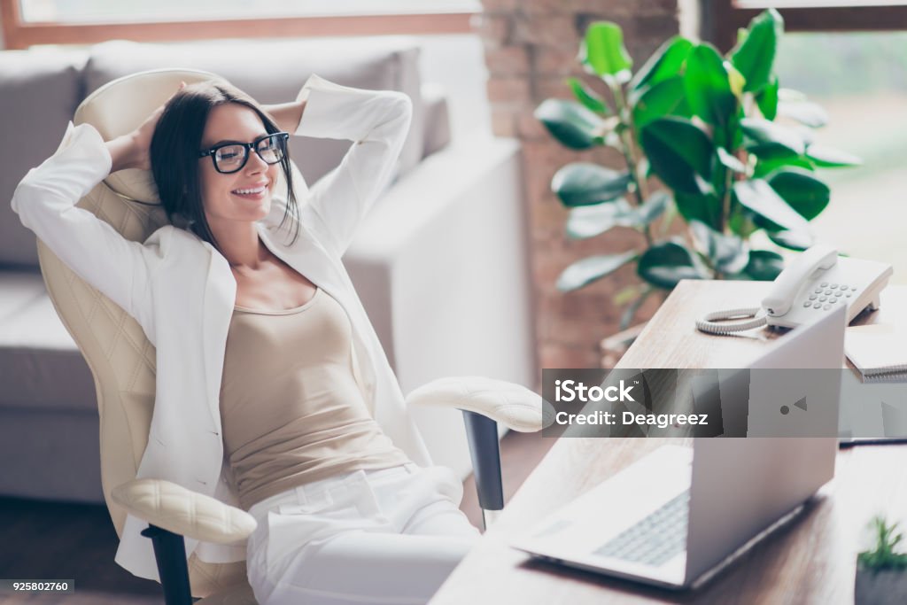 Bom, bonita e independente mulher em um terno branco, formal óculos, sentado na cadeira em sua mesa no escritório, segurando braços atrás da cabeça com os olhos perto, pensando em feriados, férias - Foto de stock de Escritório royalty-free