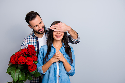 Handsome man with bristle in  shirt making  surprise for his beautiful, charming, joyful lover in casual outfit, closing her eyes with hands over grey background, women's day