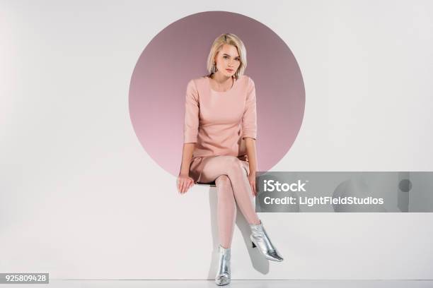 Beautiful Stylish Blonde Woman In Dress And Shiny Shoes Sitting In Hole And Looking At Camera On Grey Stock Photo - Download Image Now