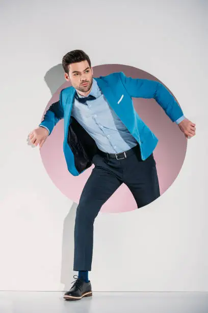 Photo of handsome stylish young man in suit and bow tie looking away while stepping through hole on grey