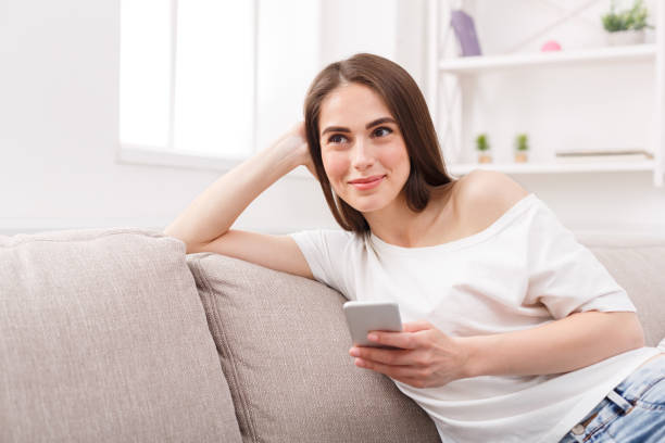 istock Smiling young woman sitting on sofa with cell phone 925833458