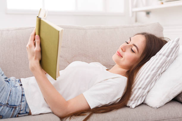 istock Happy young woman reading book on sofa 925836392
