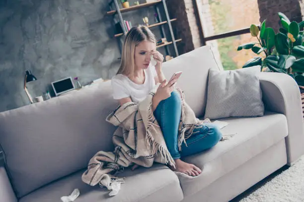 Photo of Single frustrated exhausted woman is sitting on a sofa in a living room covered with a plaid, surrounded with paper tissues, she is crying and reading old sms on her telephone