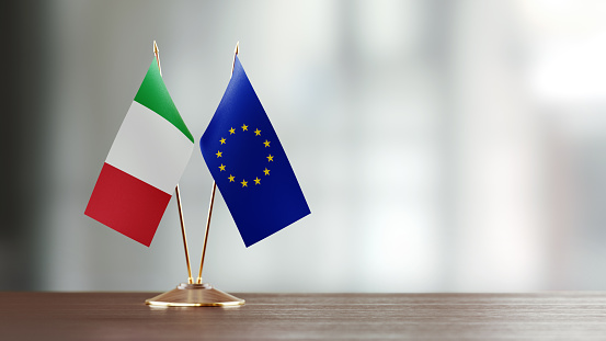 Italian and European Union flag pair on desk over defocused background. Horizontal composition with copy space and selective focus.