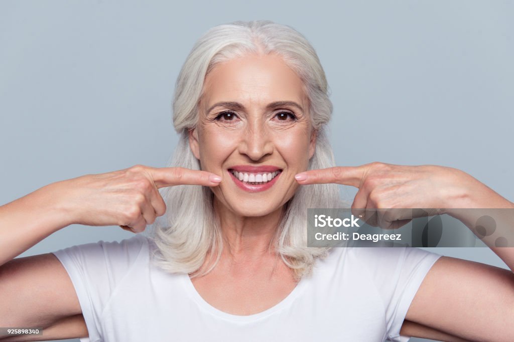 Concept of having strong healthy straight white teeth at old age. Close up portrait of happy with beaming smile female pensioner pointing on her perfect clear white teeth, isolated on gray background Dental Implant Stock Photo