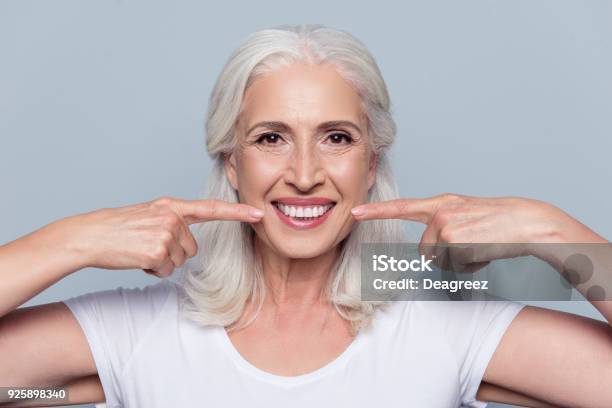 Concept Of Having Strong Healthy Straight White Teeth At Old Age Close Up Portrait Of Happy With Beaming Smile Female Pensioner Pointing On Her Perfect Clear White Teeth Isolated On Gray Background Stock Photo - Download Image Now