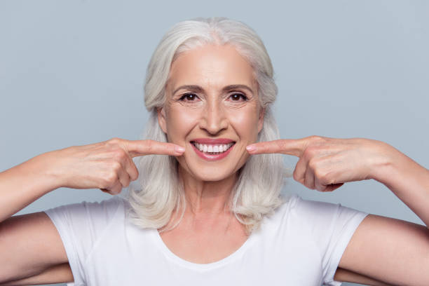 concept of having strong healthy straight white teeth at old age. close up portrait of happy with beaming smile female pensioner pointing on her perfect clear white teeth, isolated on gray background - healthy gums fotos imagens e fotografias de stock