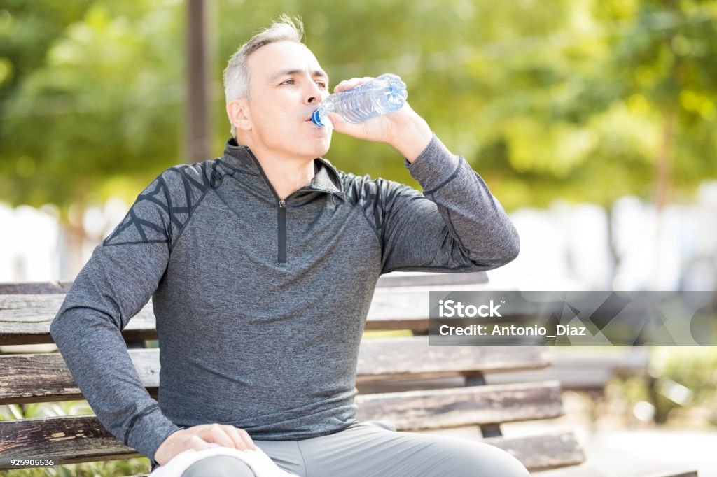 Mature man drinking water to refresh after a workout Fit mature man drinking water and taking a break from workout outdoors in a park Bench Stock Photo