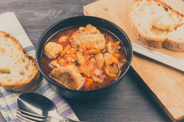 istock Braised pork with vegetables in a black bowl and a piece of bread on a rustic table 925905596