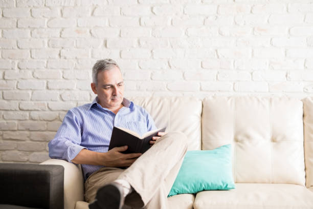 Latin man with gray hair sitting in the living room reading a novel to pass the time