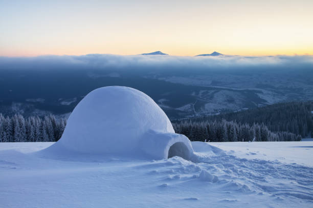 on the snowy lawn in the snowdrift there is an igloo covered by snow with the background of mountains, forests, fog and sunrise. - igloo imagens e fotografias de stock