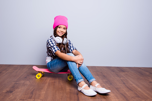Girly fashion, modern pre teens lifestyle concept. Charming attractive school girl in fashionable trendy outfit, with toothy smile, sits on bright skate on brown wooden floor, with big white headset