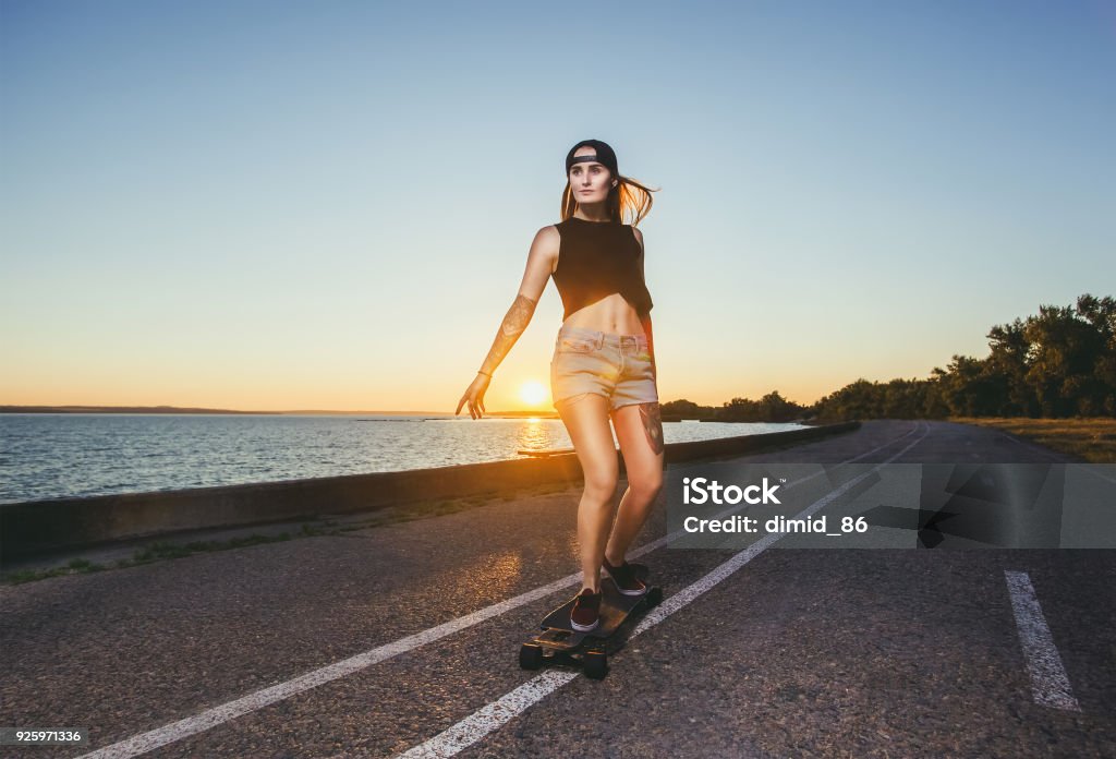 A beautiful sexy blond hipster with blue hair in a tattoo is riding a longboard A beautiful sexy blond hipster with blue hair in a tattoo is riding a longboard on an asphalt road. Longboard Skating Stock Photo