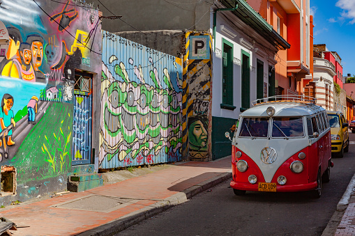 Bogota, Colombia - May 28, 2018: An old Volkswagen mini van drives through a narrow colorful street in the historic La Candelaria District in the capital city of Bogota in the South American country of Colombia. Many of the walls in the area are painted in the vibrant colours of Colombia, sometimes depicting Pre-Colombian legends or modern Street Art and Graffiti. The gate to the left is painted by the Spanish Street Artist who signs off as Pez and paints only Fish. Photo shot in the late afternoon sunlight; horizontal format. Camera: Canon EOS 5D MII. Lens: Canon EF 24-70mm F2.8L USM.