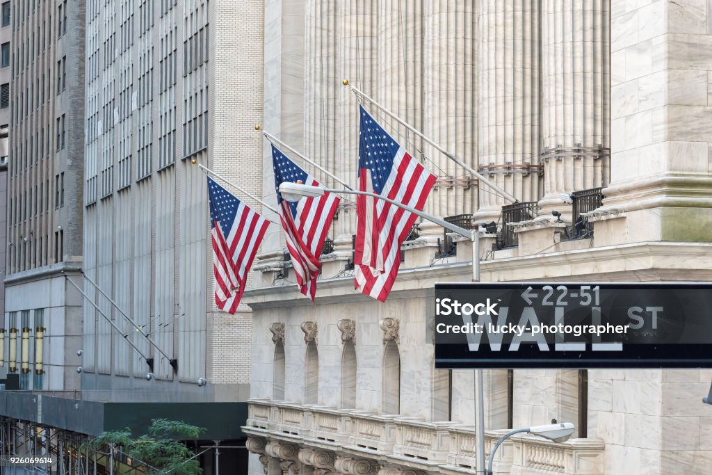 Wall street sign in New York City with New York Stock Exchange background. Wall Street sign with american flags and New York Stock Exchange in Manhattan, New York City, USA. Wall Street - Lower Manhattan Stock Photo