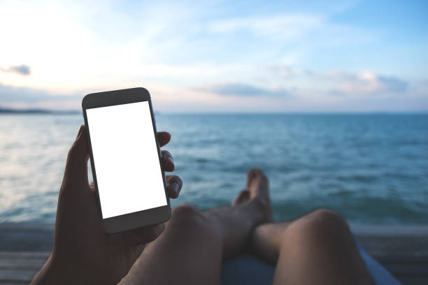 istock Mockup image of a man's hand holding white mobile phone with blank desktop screen sitting by the sea and blue sky background 926069988