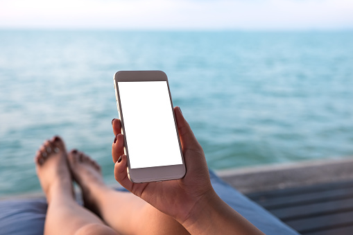 Mockup image of a woman using white mobile phone with blank desktop screen while sitting by the sea