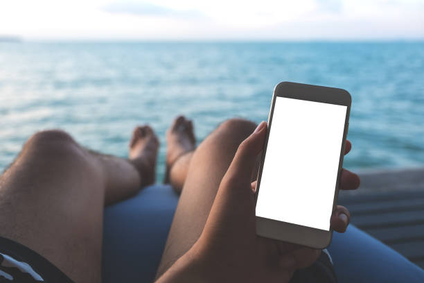 istock Mockup image of a man's hand holding white mobile phone with blank desktop screen sitting by the sea and blue sky background 926070016