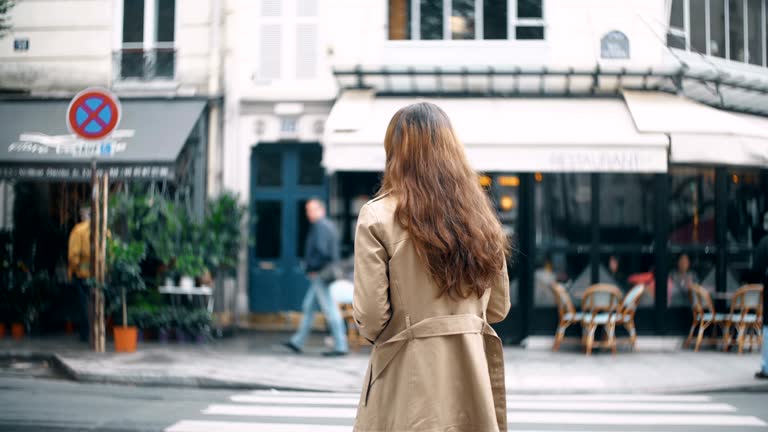 Back view of young beautiful woman crossing the road in Paris, France. Stylish female in cloak walking on the street