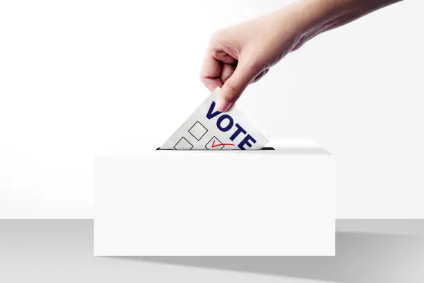 close up of hand holding voting paper for election vote into the ballot box on white background