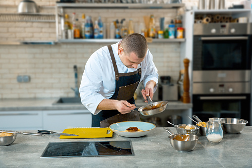 food cooking, profession and people concept - male chef cook serving plate of polenta and veal tongues with sauce at restaurant kitchen interior. cooking process