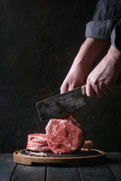 Raw tomahawk steak Man's hands cutting raw uncooked black angus beef tomahawk steaks on bones by vintage butcher cleaver on round wooden slate cutting board over dark wooden plank table. Rustic style steak vertical beef meat stock pictures, royalty-free photos & images