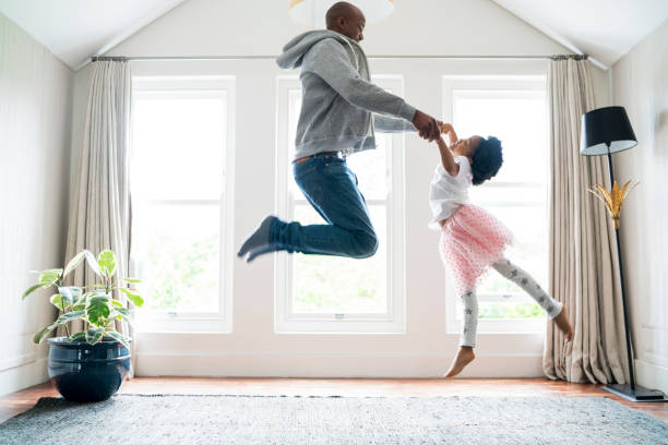 Father and daughter jumping while doing ballet Side view of father and daughter jumping while performing ballet. Man and girl are practicing at home. Family is dancing against windows. dancing dad stock pictures, royalty-free photos & images