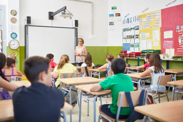Teacher teaching students in classroom at school Teacher teaching male and female students. Boys and girls learning in school. They are sitting on benches in classroom. kids using smart board stock pictures, royalty-free photos & images