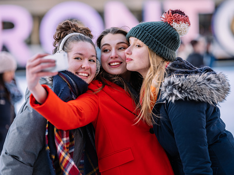 Young Caucasian teenage women taking selfies on the skating rink in downtown area of big city in North America. All 3 smiling, dressed in heavy winter clothes. In the background, unrecognizable people skating, neon signs of the city.