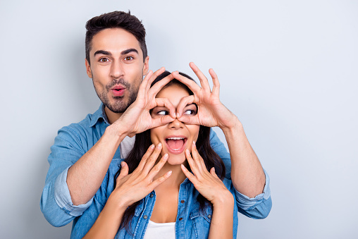 Handsome bearded man with pout lips making eyewear with fingers to his wonderful girlfriend who holding her hands on face with open mouth looking side standing over grey background