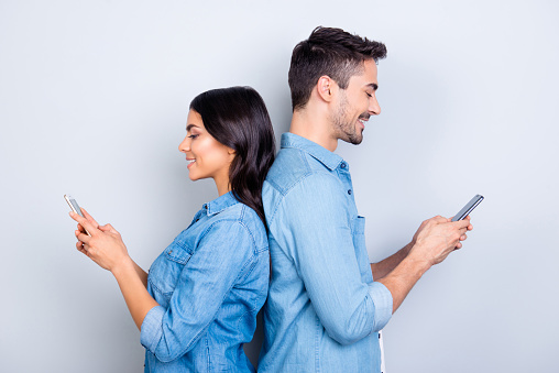 Beautiful, pretty, cute woman and handsome man in jeans shirts standing back to back and writing sms through 3g internet on smart phones over grey background