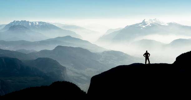 Spectacular mountain ranges silhouettes. Man reaching summit enjoying freedom. Beautiful view snow covered mountain ranges silhouettes and fog filled valleys with bright back light. South Tyrol, Itay, Alps. Happy winning success man at sunset or sunrise standing relaxed and is happy for having reached mountain top summit goal during hiking travel trek. simple living photos stock pictures, royalty-free photos & images
