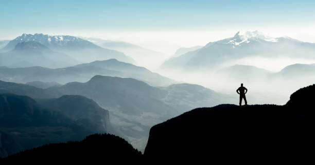 Spectacular mountain ranges silhouettes. Man reaching summit enjoying freedom. Beautiful view snow covered mountain ranges silhouettes and fog filled valleys with bright back light. South Tyrol, Itay, Alps. Happy winning success man at sunset or sunrise standing relaxed and is happy for having reached mountain top summit goal during hiking travel trek. mountain man stock pictures, royalty-free photos & images