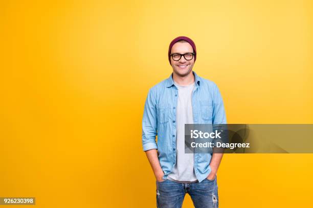 Retrato Con Espacio De Copia De Sonriente Feliz Chico En Desgaste Los Pantalones Vaqueros Gafas Gorra Roja Cogidos De La Mano En El Bolsillo Mirando A Cámara Sobre Fondo Amarillo Foto de stock y más banco de imágenes de Hombres