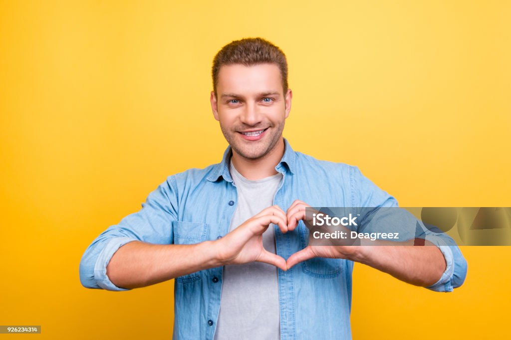 Portrait of  smiling, stunning man with stubble showing heart figure with fingers over yellow background Heart Shape Stock Photo