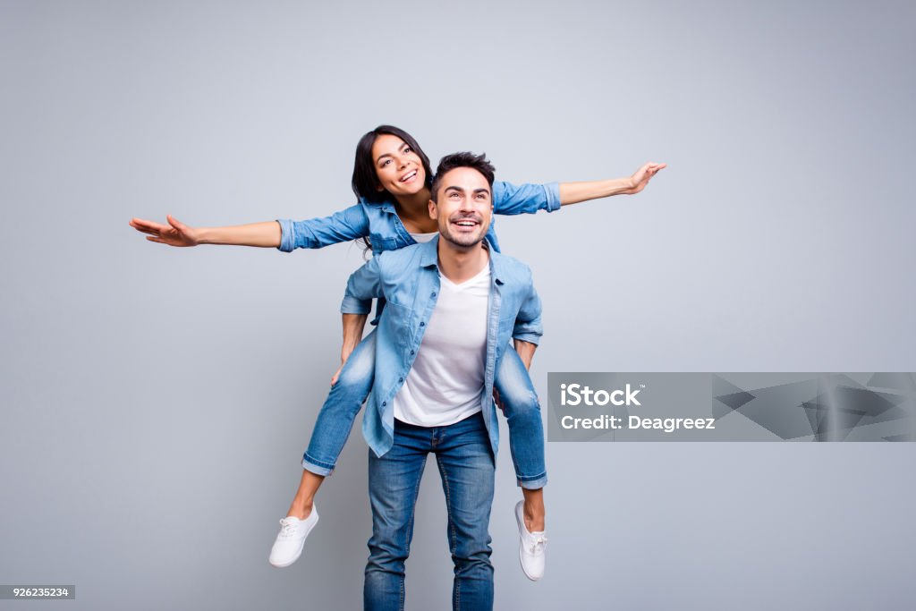 I believe I can fly. Love story of attractive, funny, cheerful couple - handsome man carrying his lover on back like plane, woman opens her hands to the side over grey background Carrying Stock Photo