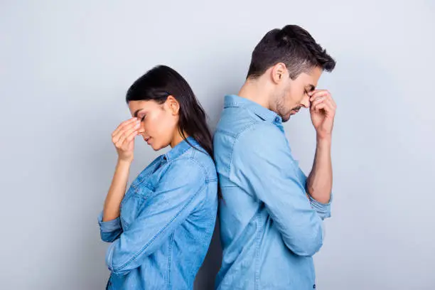 Two entrepreneurs overworked holding their fingers on nose between eyes, relationship of young family came to the dead end, standing over grey background