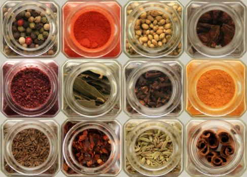 A grid arrangement of herbs and spices in glass spice jars. Each jar containing a different herb or spice.