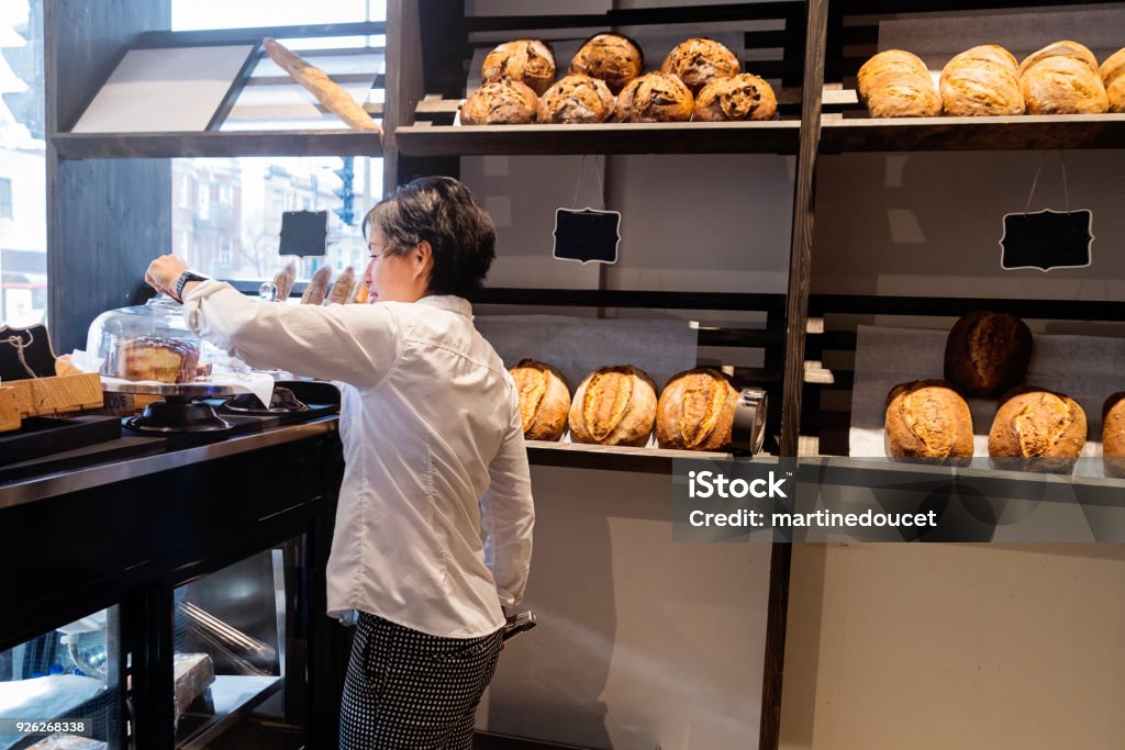 Service at the counter for employee of a small bakery shop. Employee of a small local bakery shop getting a pastry behind the counter. Horizontal waist up indoors shot with copy space. This was taken in Montreal, Canada. 50-54 Years Stock Photo