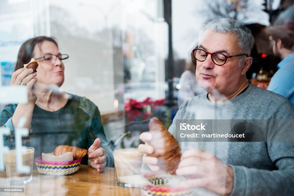Senior couple eating in a small local bakery shop. Senior couple eating in a small local bakery shop. This was shot through the window from the street. Focus on the man’s face. Horizontal waist up indoors shot with copy space. This was taken in Montreal, Canada. Eating Stock Photo