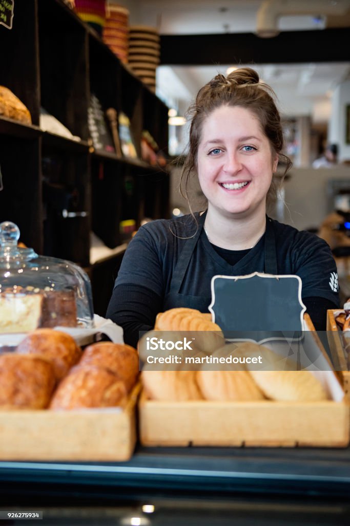 Customer service in a small local bakery shop. Customer service in a small local bakery shop. Portrait of employee standing behind the counter. Vertical waist up indoors shot with copy space. This was taken in Montreal, Canada. Bakery Stock Photo