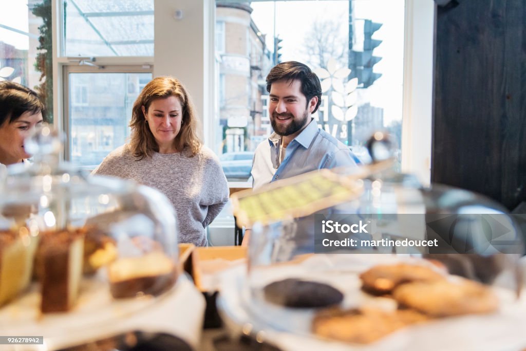 Customer choosing something to eat in a small local bakery shop. Customer choosing something to eat in a small local bakery shop. Focus on faces of clients. Pastries and cookies in the foreground. Horizontal waist up indoors shot with copy space. This was taken in Montreal, Canada. Bakery Stock Photo