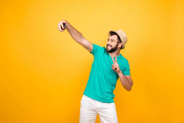 Photo of Hey what's up? Happy funky cheerful joyful man clothed in casual outfit taking a self portrait on his new smartphone and showing two fingers, copyspace