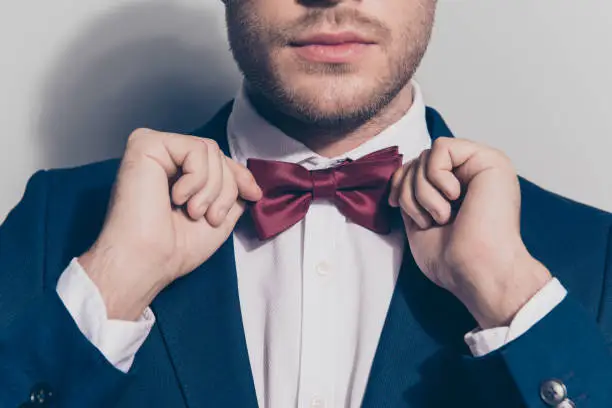 Photo of Close up portrait of half face - bearded stubble man ties a bowtie at the collar, correcting red bow on his white shirt over grey background
