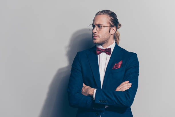 ritratto di elegante uomo barbuto in occhiali, giacca blu, camicia bianca con papillon rosso, quadrato tascabile e coda in piedi con le mani incrociate, viso mezzo rivolto a lato su sfondo grigio - pocket suit glasses jacket foto e immagini stock