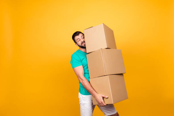Concept of courier and messenger service. Grimacing handsome guy is trying to keep big stack of cardboard bosex in his hand, isolated on yellow background