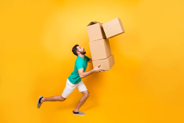 istock Funny nervous man is carrying his stuff in boxes to the recently bought flat. He is holding cardboard boxes and one is falling down, isolated on yellow background 926314222